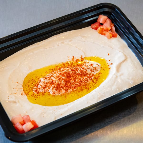 Creamy hummus topped with olive oil, paprika, and diced tomatoes in a black container.