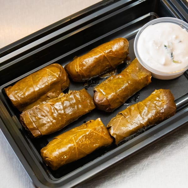 Six dolmades, or stuffed grape leaves, with a side of white dipping sauce in a black tray.
